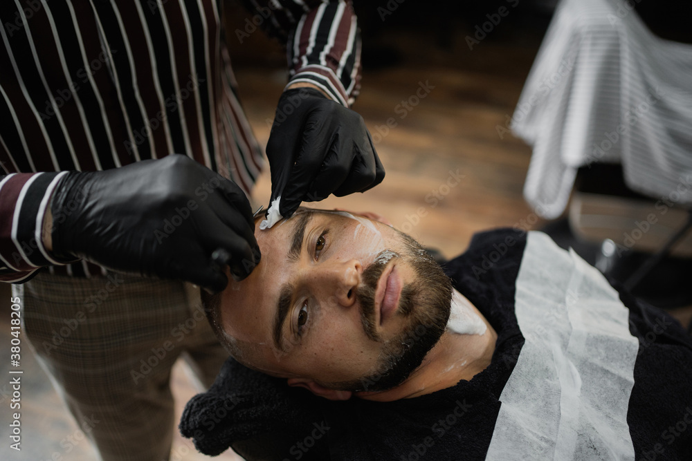 Fototapeta premium young man confident brutal Caucasian appearance, doing a haircut of his beard and hair on his head. dangerous razor blade shaving foam
