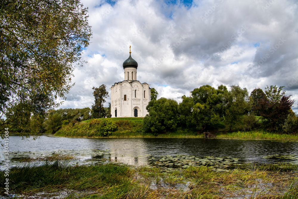 snow-white stone old church against the background of a green meadow and lake before a summer thunderstorm