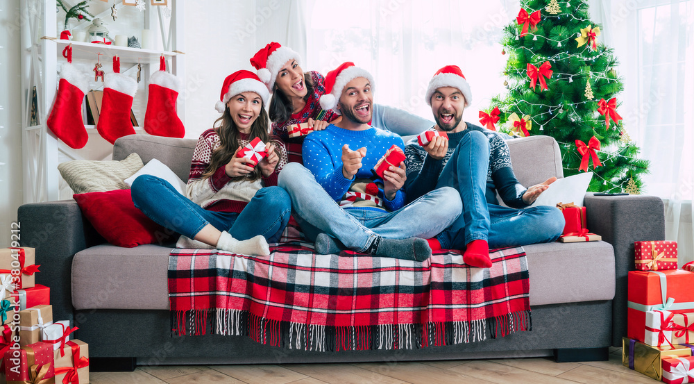 Merry Christmas and Happy New Year. The big happy excited group of young friends in Christmas santa hats with gift boxes are having fun together while sitting on the couch. Merry Christmas and Happy New Year. The big happy excited group of young friends in Christmas santa hats with gift boxes are having fun together while sitting on the couch.