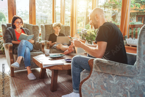 Foto Mother, father and son family sitting together at Sunroom in cozy armchairs and reading books, using laptop or browsing a smartphone