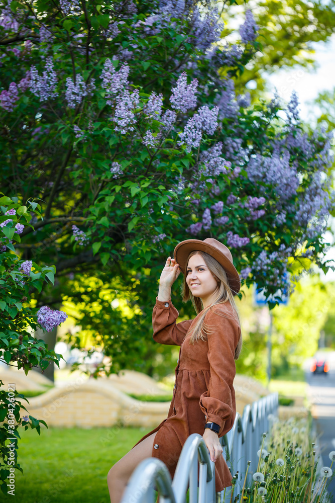 Naklejka premium Closeup young girl standing on a background of lilac bushes. she is of European appearance in a brown hat and dress