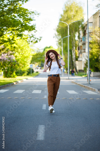 A beautiful girl in a brown hat and a white T-shirt is walking on the road on a warm sunny day. Beautiful young woman in a hat with a smile on her face