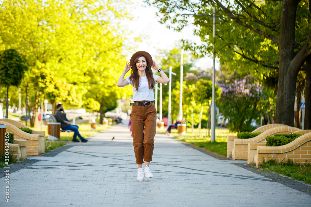 Young happy woman in a hat walks along an alley in a park. A girl of European appearance with a smile on her face on a bright sunny summer day Young happy woman in a hat walks along an alley in a park. A girl of European appearance with a smile on her face on a bright sunny summer day