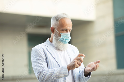 Coronavirus. Cleaning hands with sanitizer spray in city. Bearded senior man wearing in medical protective mask on street. Sanitizer to prevent Coronavirus, Covid-19, flu.Virus and illness protection.