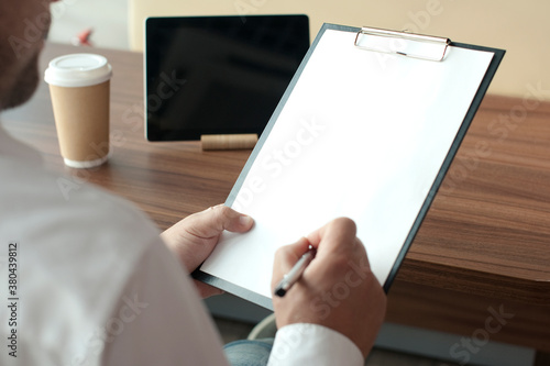 Businessman in a white shirt with a digital tablet in his hands signs a contract in the office. Workplace with a cup of coffee and a document with a pen on a wooden table.