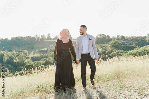 Muslim love story of mixed couple. Man and woman smiles and walks on the green hills . Woman weared in hijab looks to her man. Advert for on-line dating agency