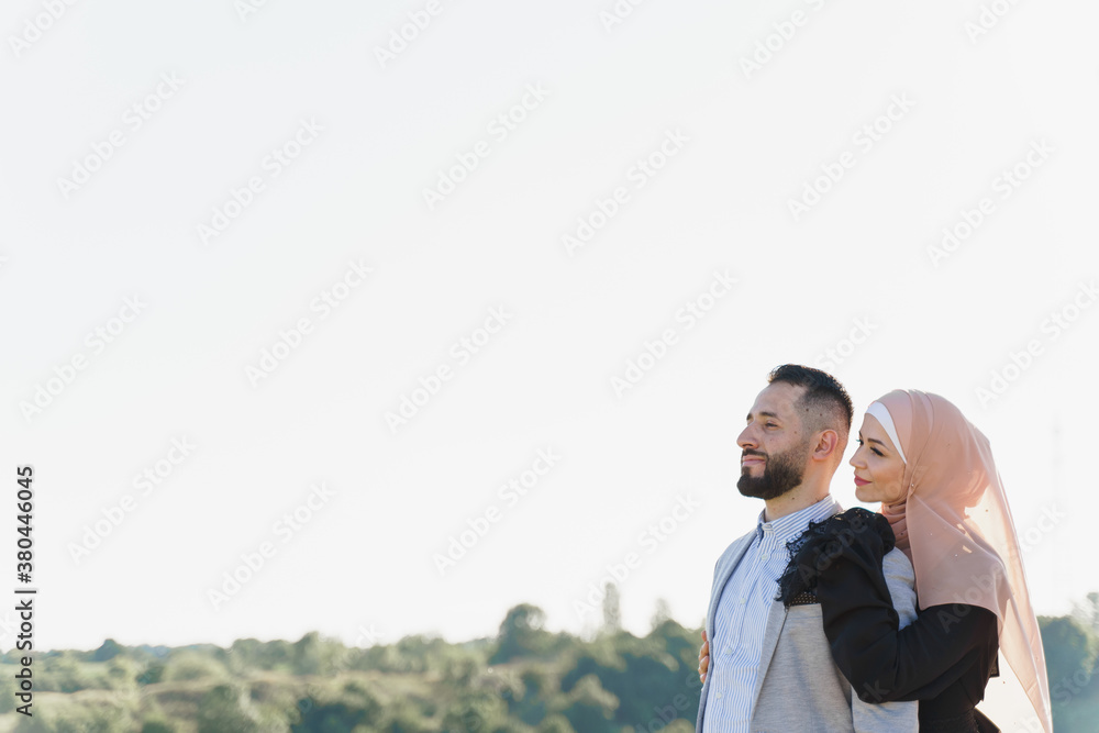 Muslim love story. Mixed couple smiles and hugs on the green hills . Woman weared in hijab looks to her man. Advert for on-line dating agency Muslim love story. Mixed couple smiles and hugs on the green hills . Woman weared in hijab looks to her man. Advert for on-line dating agency