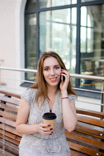Beautiful woman is speaking on the phone with a cup of coffee in the hand, young girl is sitting on a bench against the glass facade in the city