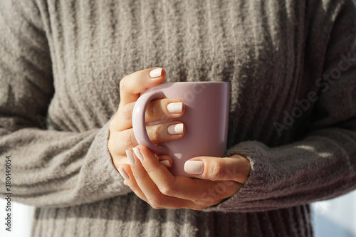 beautiful female hands with well-groomed white manicure are holding a mug of hot drink. The position of the hands is frontal, against a background of a gray beige knitted cashmere sweater with a verti photo