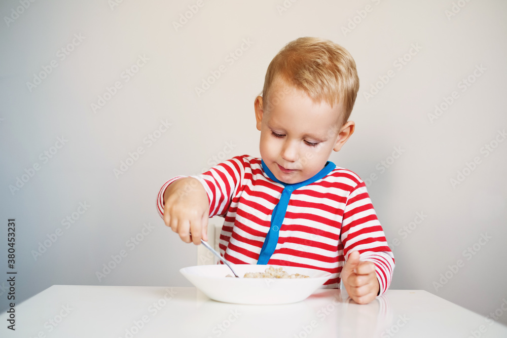 child is holding a spoon in his hands and eats porridge while sitting at the table. child is holding a spoon in his hands and eats porridge while sitting at the table.