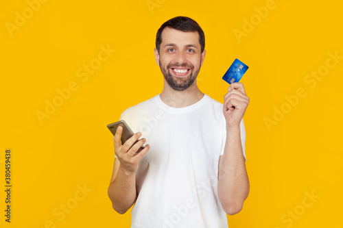 Young man with a beard in a white t-shirt holding a credit card. by credit card to pay online using a smartphone. Stands on isolated yellow background