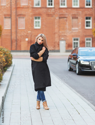Outdoors lifestyle fashion portrait of stunning blonde young woman walking on the street. Wrapping up in a coat from the cold. Wearing stylish black coat and ankle boots. Cold weather. Feeling cold © Olesya Kuprina