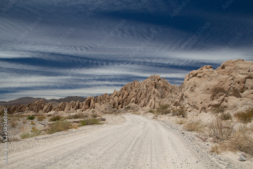 Fototapeta premium Road trip. Driving along the dirt road across the arid desert. The route, rocky valley and beige sandstone hills under a beautiful blue sky.