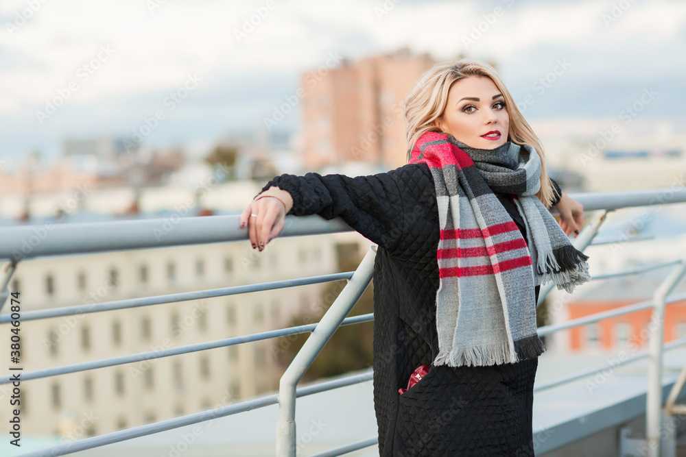 Outdoors lifestyle fashion portrait of stunning blonde girl. Posing on the roof of old town. Dreaming, enjoying life. Wearing stylish black oversized coat and wide scarf. Cold weather. Romantic mood.
