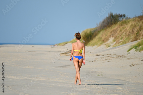 A young woman walks along a deserted beach. The summer sun warms her back, on which a mobile phone is fixed with a bra strap.