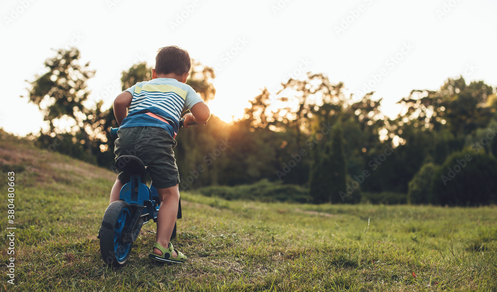 Back view photo of a caucasian boy riding the bike in a park on the grass during a beautiful sunset Back view photo of a caucasian boy riding the bike in a park on the grass during a beautiful sunset