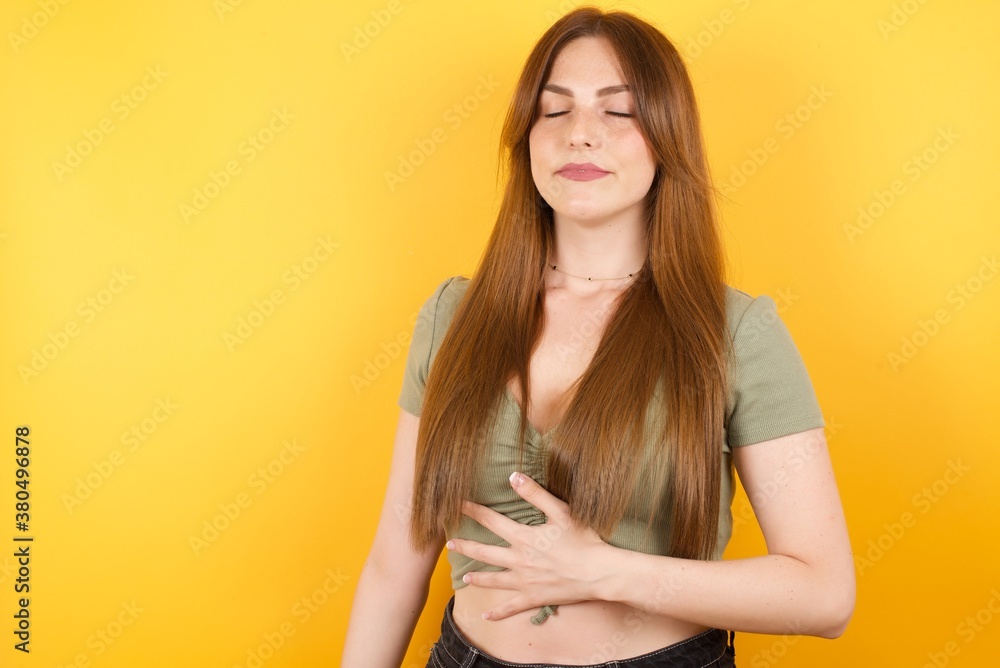 Young caucasian woman with long hair wearing green tshirt standing over isolated yellow background touches tummy, smiles gently, eating and satisfaction concept. Young caucasian woman with long hair wearing green tshirt standing over isolated yellow background touches tummy, smiles gently, eating and satisfaction concept.