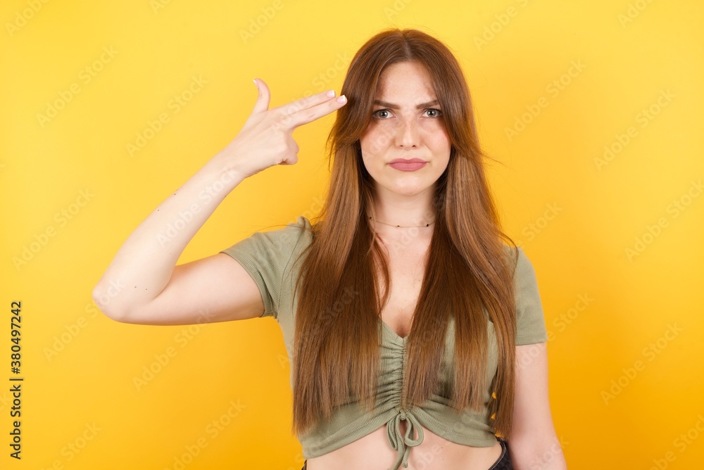Unhappy Young caucasian woman with long hair wearing green tshirt standing over isolated yellow background imitates gun shoot makes suicide gesture keeps two fingers on temples has puzzled expression Unhappy Young caucasian woman with long hair wearing green tshirt standing over isolated yellow background imitates gun shoot makes suicide gesture keeps two fingers on temples has puzzled expression