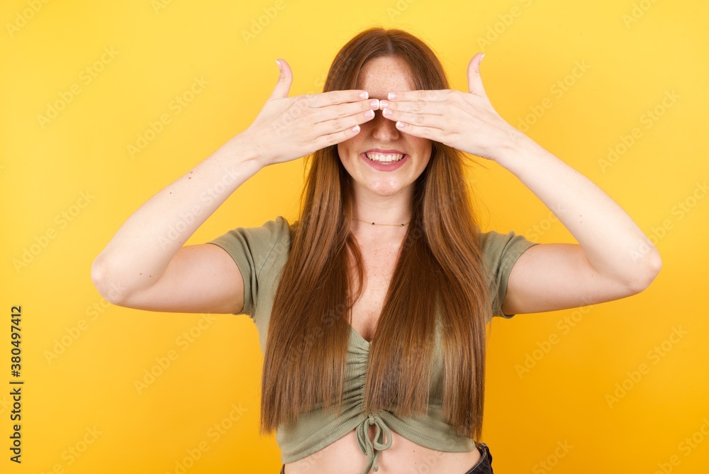 Young caucasian woman with long hair wearing green tshirt standing over isolated yellow background covering eyes with hands smiling cheerful and funny. Blind concept. Young caucasian woman with long hair wearing green tshirt standing over isolated yellow background covering eyes with hands smiling cheerful and funny. Blind concept.