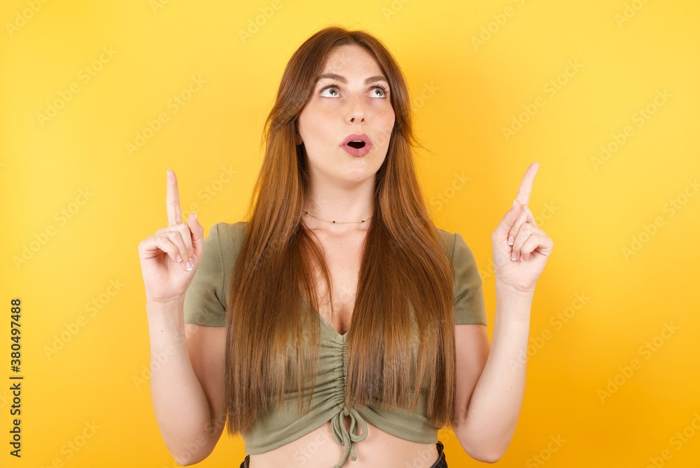 Young caucasian woman with long hair wearing green tshirt standing over isolated yellow background amazed and surprised looking up and pointing with fingers and raised arms. Young caucasian woman with long hair wearing green tshirt standing over isolated yellow background amazed and surprised looking up and pointing with fingers and raised arms.