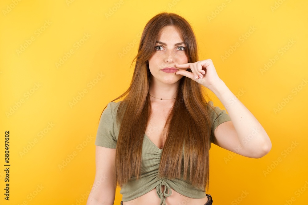 Young caucasian woman with long hair wearing green tshirt standing over isolated yellow background mouth and lips shut as zip with fingers. Secret and silent, taboo talking. Young caucasian woman with long hair wearing green tshirt standing over isolated yellow background mouth and lips shut as zip with fingers. Secret and silent, taboo talking.