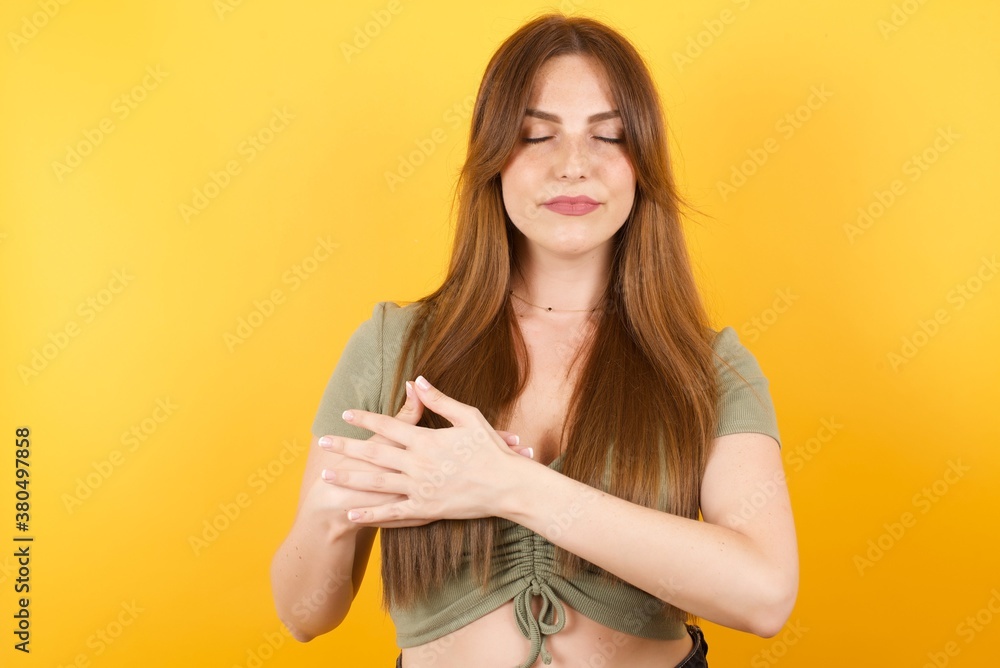 Young caucasian woman with long hair wearing green tshirt standing over isolated yellow background smiling with hands on chest with closed eyes and grateful gesture on face. Health concept. Young caucasian woman with long hair wearing green tshirt standing over isolated yellow background smiling with hands on chest with closed eyes and grateful gesture on face. Health concept.