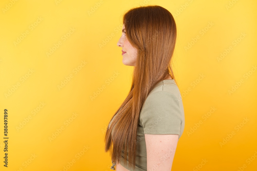 Young caucasian woman with long hair wearing green tshirt standing over isolated yellow background looking to side, relax profile pose with natural face with confident smile. Young caucasian woman with long hair wearing green tshirt standing over isolated yellow background looking to side, relax profile pose with natural face with confident smile.