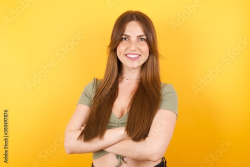 Young caucasian woman with long hair wearing green tshirt standing over isolated yellow background happy face smiling with crossed arms looking at the camera. Positive person.