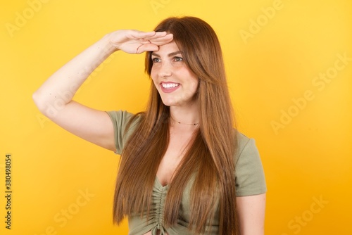 Young caucasian woman with long hair wearing green tshirt standing over isolated yellow background very happy and smiling looking far away with hand over head. Searching concept.