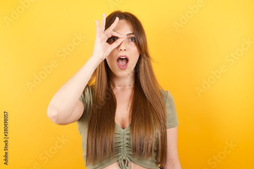 Young caucasian woman with long hair wearing green tshirt standing over isolated yellow background doing ok gesture shocked with surprised face, eye looking through fingers. Unbelieving expression. © Roquillo