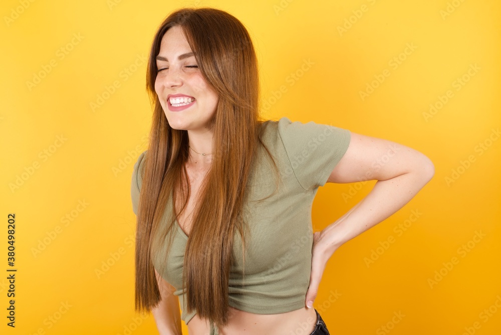 Fototapeta premium Young caucasian woman with long hair wearing green tshirt standing over isolated yellow background Suffering of backache, touching back with hand, muscular pain