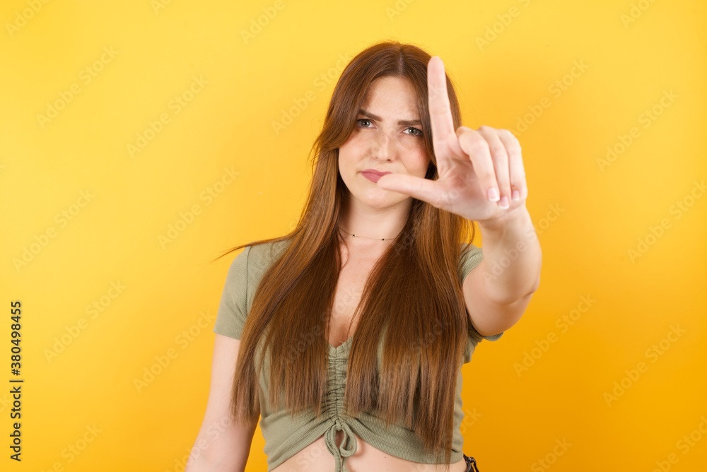 Young caucasian girl wearing sexy t-shirt over isolated yellow background making fun of people with fingers on forehead doing loser gesture mocking and insulting. Young caucasian girl wearing sexy t-shirt over isolated yellow background making fun of people with fingers on forehead doing loser gesture mocking and insulting.