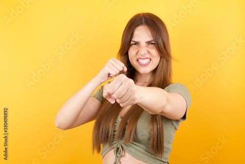 Bild auf Leinwand Portrait of strong and determined caucasian busineswoman in red shirt studio shot isolated on yellow background punching air with fist and looking confidently at camera,  fighting spirit