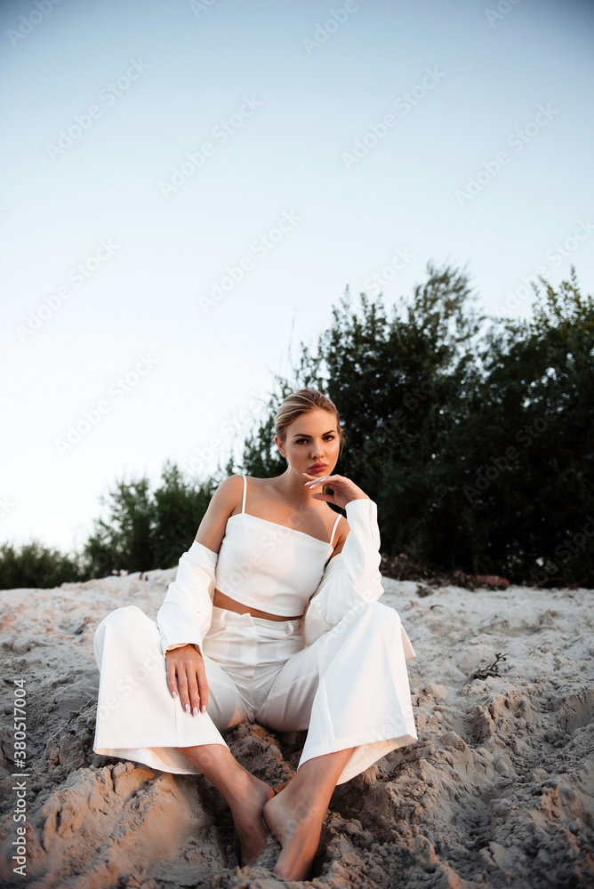 Effective blonde in white suit sitting on sand with barefoot