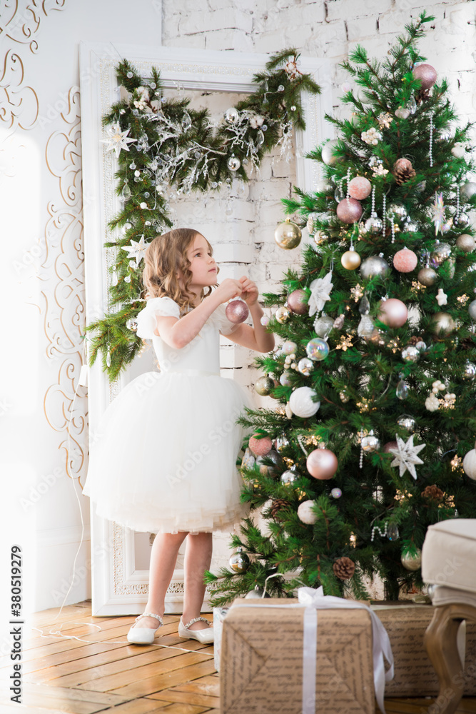a beautiful little girl in a white dress is decorating a Christmas tree. home decoration for new year celebration. winter vacation. happy childhood. a beautiful little girl in a white dress is decorating a Christmas tree. home decoration for new year celebration. winter vacation. happy childhood.