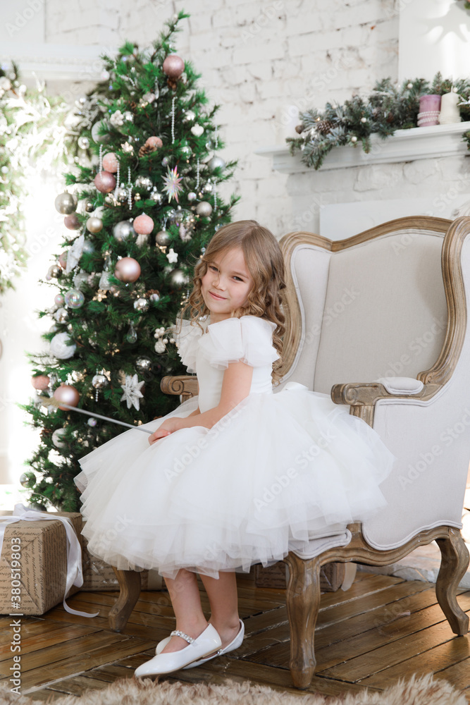 little beautiful girl in a white fluffy, elegant dress with magic wands sitting in a chair on the background of Christmas trees in the interior of the loft. new year holidays and vacations. little beautiful girl in a white fluffy, elegant dress with magic wands sitting in a chair on the background of Christmas trees in the interior of the loft. new year holidays and vacations.