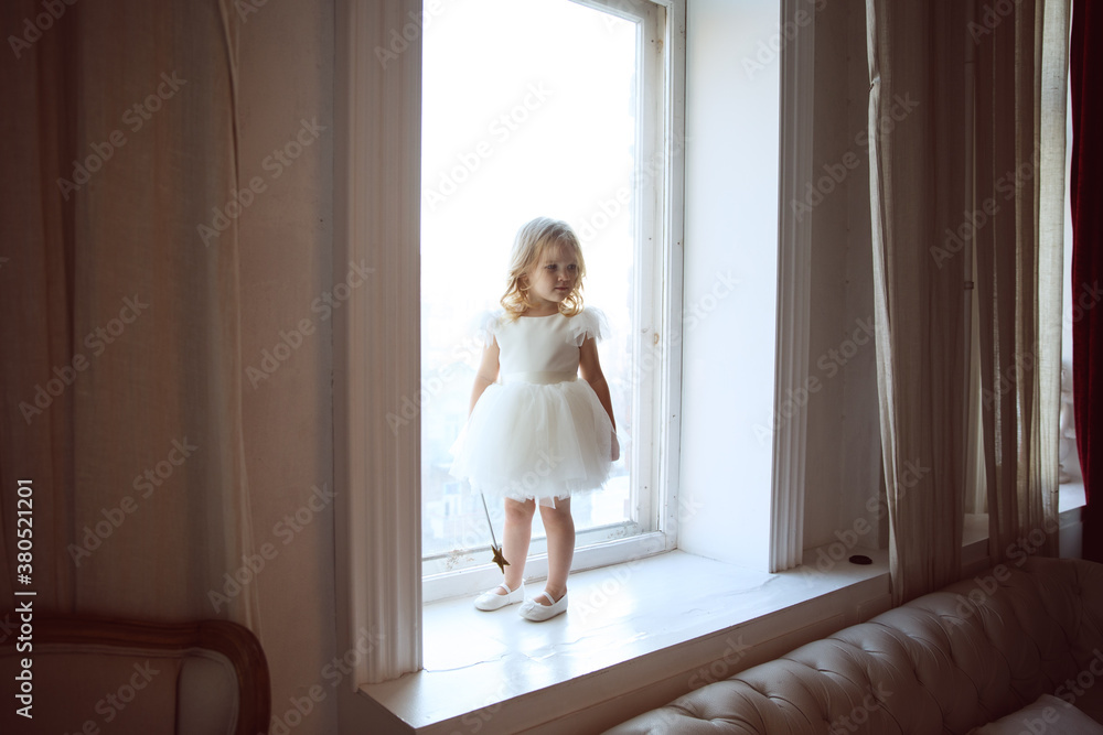 a little girl in a smart, fluffy white dress with a magic wand in her hands near the window and waits for Santa Claus or the tooth fairy. children's fairy tales and dreams. new year holiday. a little girl in a smart, fluffy white dress with a magic wand in her hands near the window and waits for Santa Claus or the tooth fairy. children's fairy tales and dreams. new year holiday.