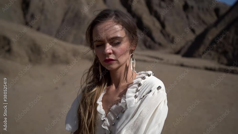 A young female tourist in a light dress stands against the background of mountains in a sand pit and poses, the wind blows her hair. A young female tourist in a light dress stands against the background of mountains in a sand pit and poses, the wind blows her hair.