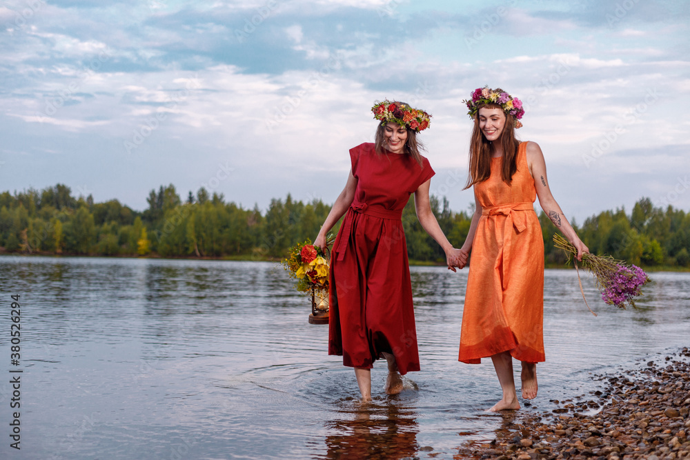 Fototapeta premium Two young and lovely girls in flower wreaths in nature. Ancient pagan origin celebration concept. Summer solstice day. Mid summer. Ancient rituals.