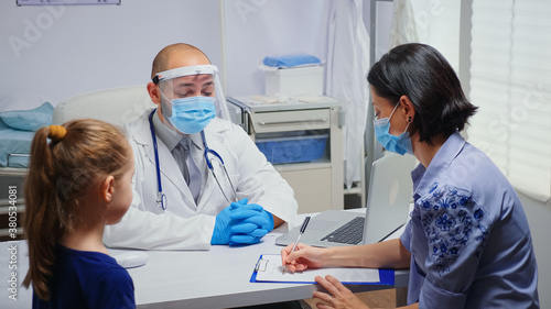 Mother writing on clipboard doctor instructions wearing protection mask. Physician specialist in medicine providing health care services consultation treatment examination in hospital during covid-19