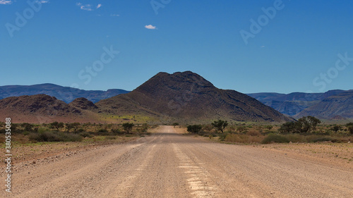 Gravel road leading towards a mountain with slightly green grass and some trees shortly after the end of rainy season near Sesriem, Namib desert, Namibia, Africa.