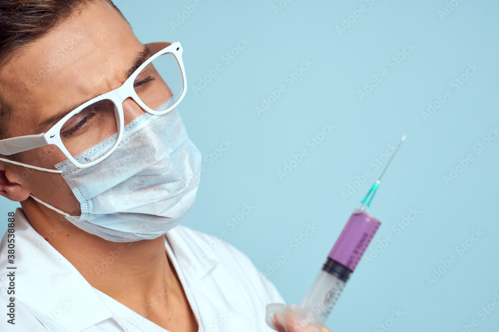 Doctor holds a syringe in his hand with a red liquid on a blue background medical gown and a protective mask Doctor holds a syringe in his hand with a red liquid on a blue background medical gown and a protective mask