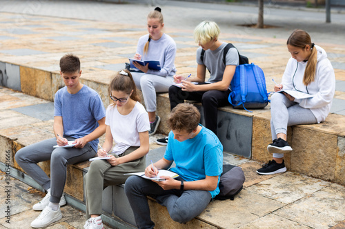 Diligent teenagers preparing schoolwork, sitting on steps outside school in sunny spring day.