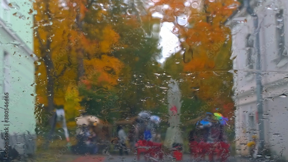 Blurred abstract image of autumn city street, apartment building, orange trees through the glass with raindrops. Rainy sad overcast weather. Cityscape through the window. Water drops.