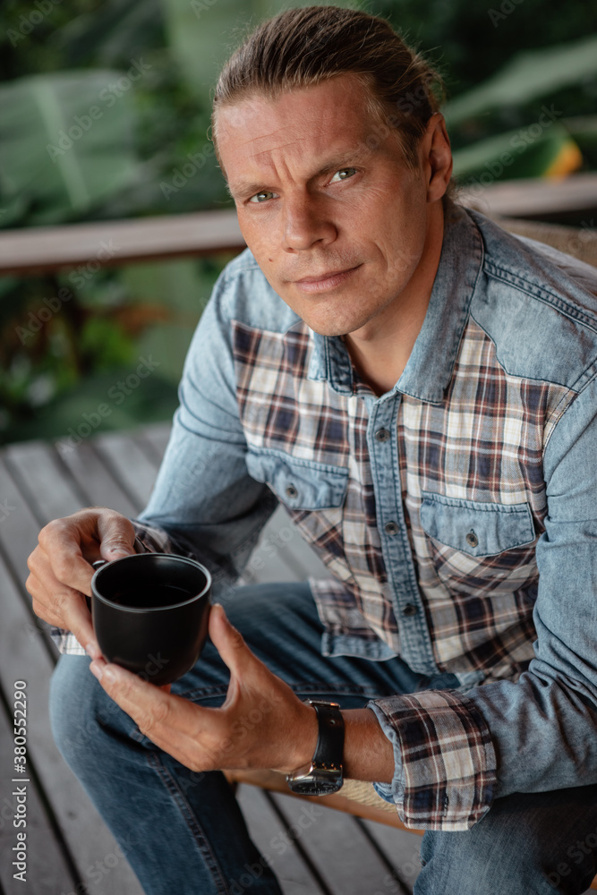 Handsome young man with long hair in checkered shirt hold in hands cup of coffee, look at the camera Handsome young man with long hair in checkered shirt hold in hands cup of coffee, look at the camera
