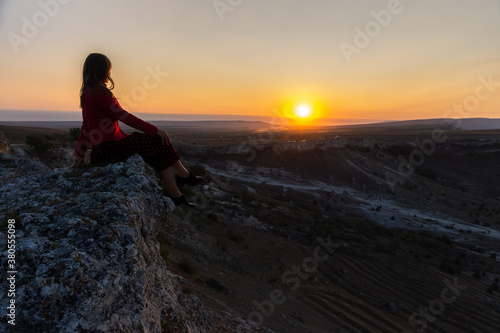 The girl sits on the edge of the cliff and looks at the sun. Beautiful dark-haired girl posing in a dress against the background of mountains. Dawn in the mountains. A girl alone with nature.