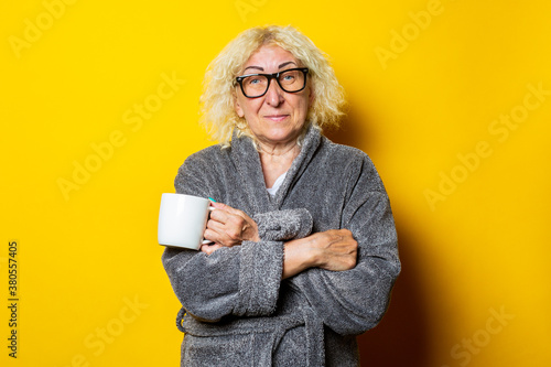 Smiling old woman in gray bathrobe holding a cup with coffee on yellow background.