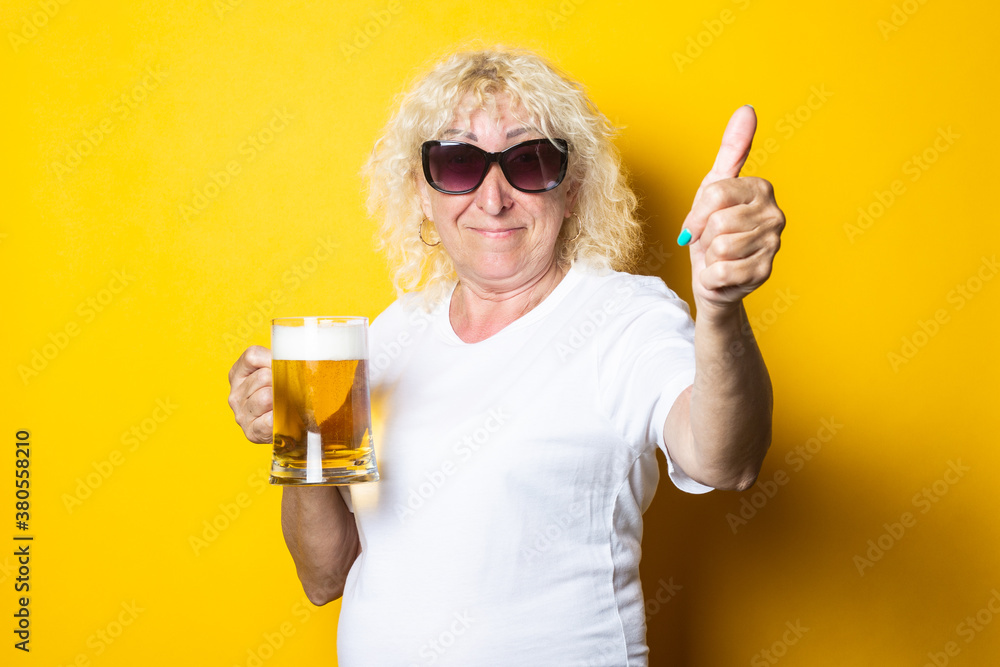 Smiling blonde old woman in sunglasses holds a glass of beer and shows a gesture class on a yellow background Smiling blonde old woman in sunglasses holds a glass of beer and shows a gesture class on a yellow background