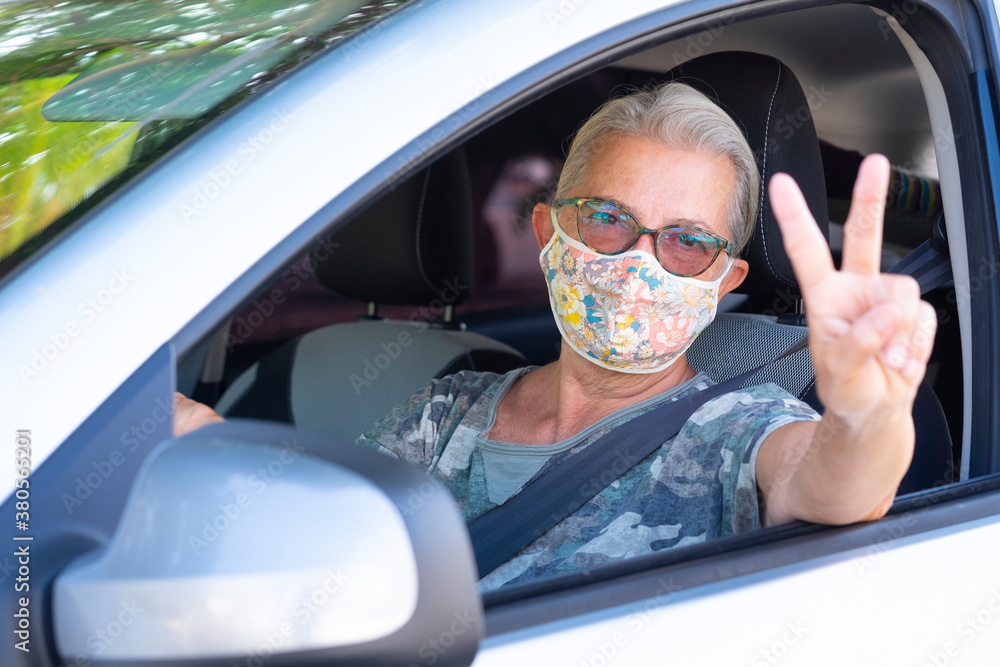 Naklejka premium Attractive senior woman with white hair wearing surgical mask gesturing ok with hand while driving her car