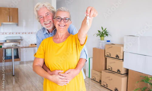 Happy smiling couple of senior people white hair hugging  holding the keys of the new empty apartment with moving boxes on the floor - concept of active elderly people and new beginning like retired photo